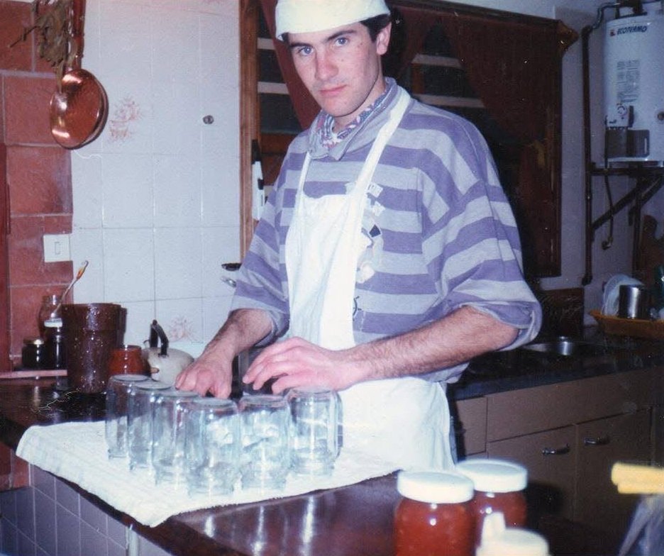 Foto antigua: un joven con gorro y delantal blanco acomoda frascos de vidrio sobre la mesada de una cocina